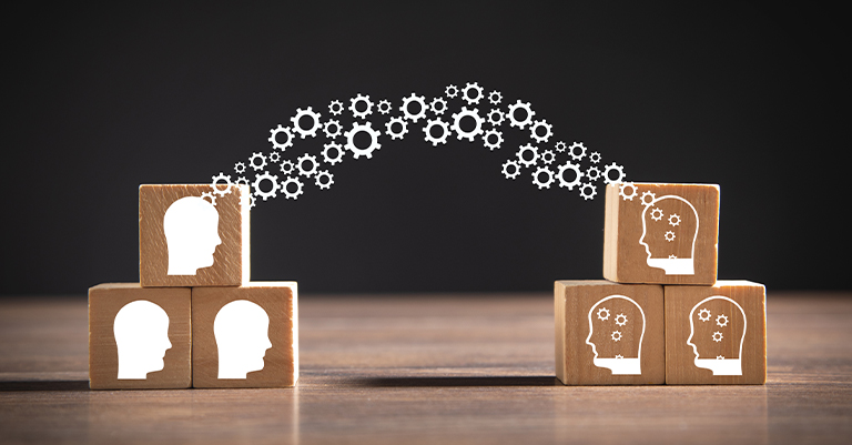 TransferLearn_1 • Andrew Roche AI Wooden blocks with human head icons connected by gear symbols, representing the transfer of knowledge and ideas.