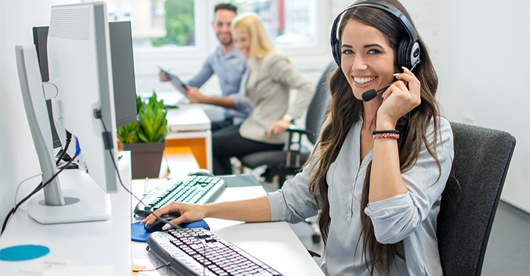 TextAnalysis_4 • Andrew Roche AI Smiling female customer service representative wearing a headset, working at her computer, with two colleagues chatting in the background.
